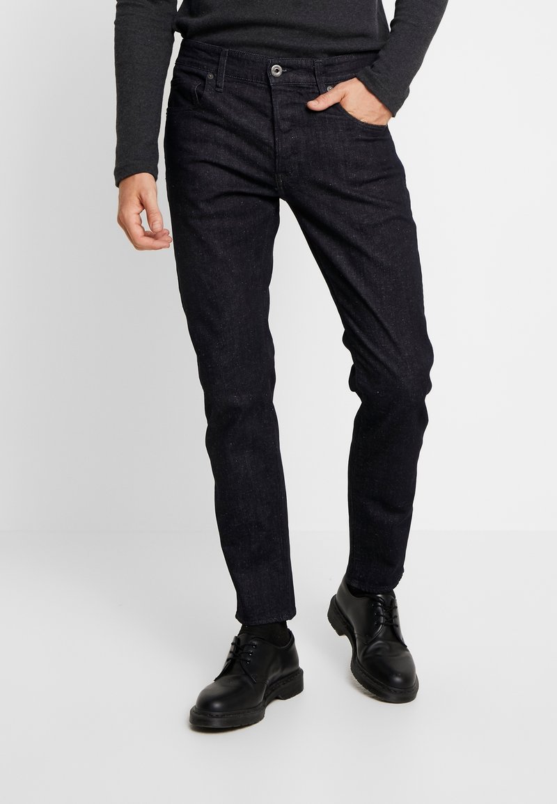 Jeans de mezclilla oscura con un corte ajustado, que cuentan con cinco bolsillos, cierre de botón y una ligera textura en la superficie. Combinados con zapatos negros de cordones.