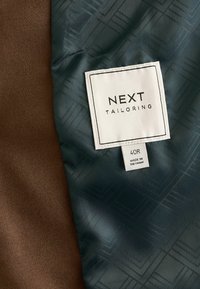 Zeleni uzorčasti materijal s sjajem pored smeđeg teksturiranog materijala. Oznaka prikazuje "NEXT TAILORING" i "40R" uz "MADE IN VIETNAM."