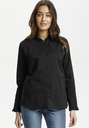 Camicia nera con bottoni realizzata in tessuto morbido, con colletto e polsini arricciati, dal taglio relaxed. Indossata con jeans blu.