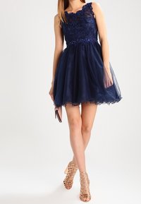 Mörkblå cocktailklänning med en figursydd spetsliv, dekorerad midja och en voluminös tyllkjol. Har justerbara axelremmar och intrikata blommönster.