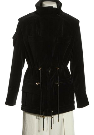 Schwarze Jacke mit langen Ärmeln, hohem Kragen, Frontreißverschluss und Kordelzug in der Taille, mit mehreren Taschen, auf einem Schaufensterpuppen-Torso präsentiert.