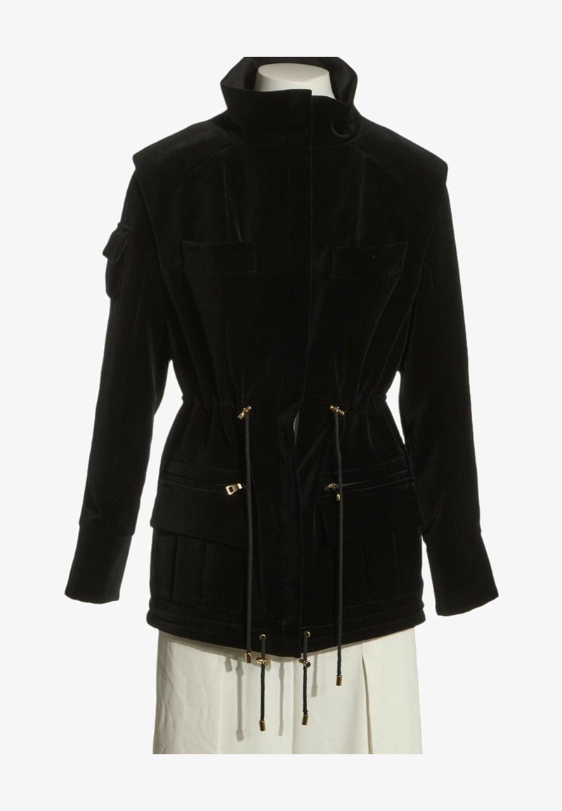 Veste noire à manches longues avec col montant, fermeture éclair devant et taille à cordon, plusieurs poches, présentée sur un torse de mannequin.