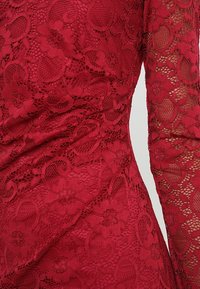 Robe rouge en dentelle avec motifs floraux, manches longues et surface texturée, mettant en valeur une broderie délicate et une transparence.