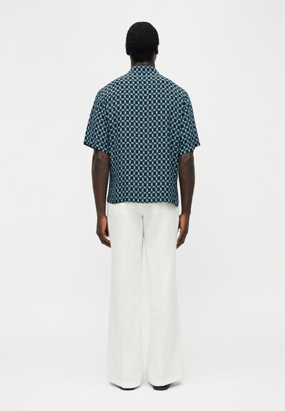 Άνδρας που στέκεται με την πλάτη, φοράει μαύρο κοντομάνικο πουκάμισο με μπλε γεωμετρικά σχέδια και φαρδιά λευκή παντελόνα.