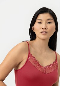 Camisole rouge avec un décolleté en dentelle florale transparente, des fines bretelles et une coupe ajustée. Tissu lisse et doux avec un léger éclat.