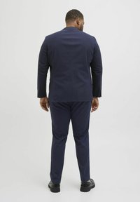 Costume bleu marine avec une coupe ajustée, tissu texturé et une fente simple au dos. Associé à des chaussures noires, vu de l'arrière.