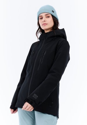 Zwarte waterdichte jas met een capuchon, ritszakken en verstelbare manchetten. Gedragen met een lichtblauwe geribbelde beanie.