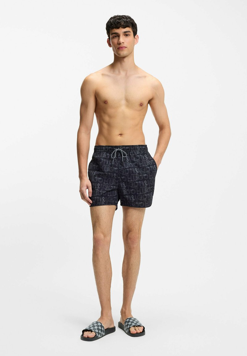 Czarne kąpielówki w wzory z sznurkiem w talii, z logo marki powtarzającym się w designie. Sparowane z wzorzystymi klapkami na suwak.