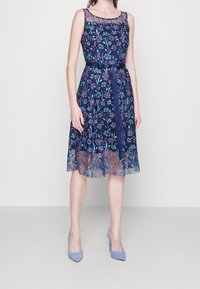 Robe bleu marine brodée de fleurs avec encolure en tulle et ruban à la taille, dotée d'une jupe évasée à hauteur des genoux et d'un tissu légèrement texturé.