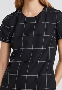 Svart kortärmad topp med vit rutmönster. Rund halsringning, slät struktur. Har en subtil sidoficka för praktisk användning.