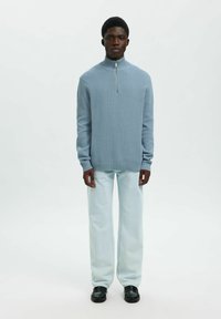 Mladic stoji odjeven u svijetloplavi pulover s polu-zipom, svijetloplave široke traperice i crne mokasine na bijeloj pozadini.