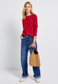 Röd rippad långärmad topp med knappdetaljer vid ärmsluten, ihopparad med lösa blå denimjeans och en beige mockaväska. Vita sneakers kompletterar outfiten.