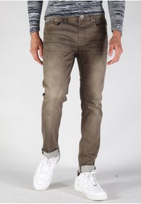 Vaqueros de mezclilla marrones con corte slim, que incluyen un cierre de cremallera, bolsillos frontales y dobladillos enrollados, combinados con zapatillas blancas.