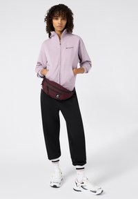Sudadera de cremallera de color púrpura claro, pantalones holgados negros, riñonera color burdeos y zapatillas blancas con detalles rosas. Pelo rizado peinado de forma natural.
