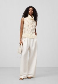Crème mouwloze top met textuur en gelaagd fabric, hoge kraag, en crème wijde broek. Model houdt een kleine crème handtas vast.