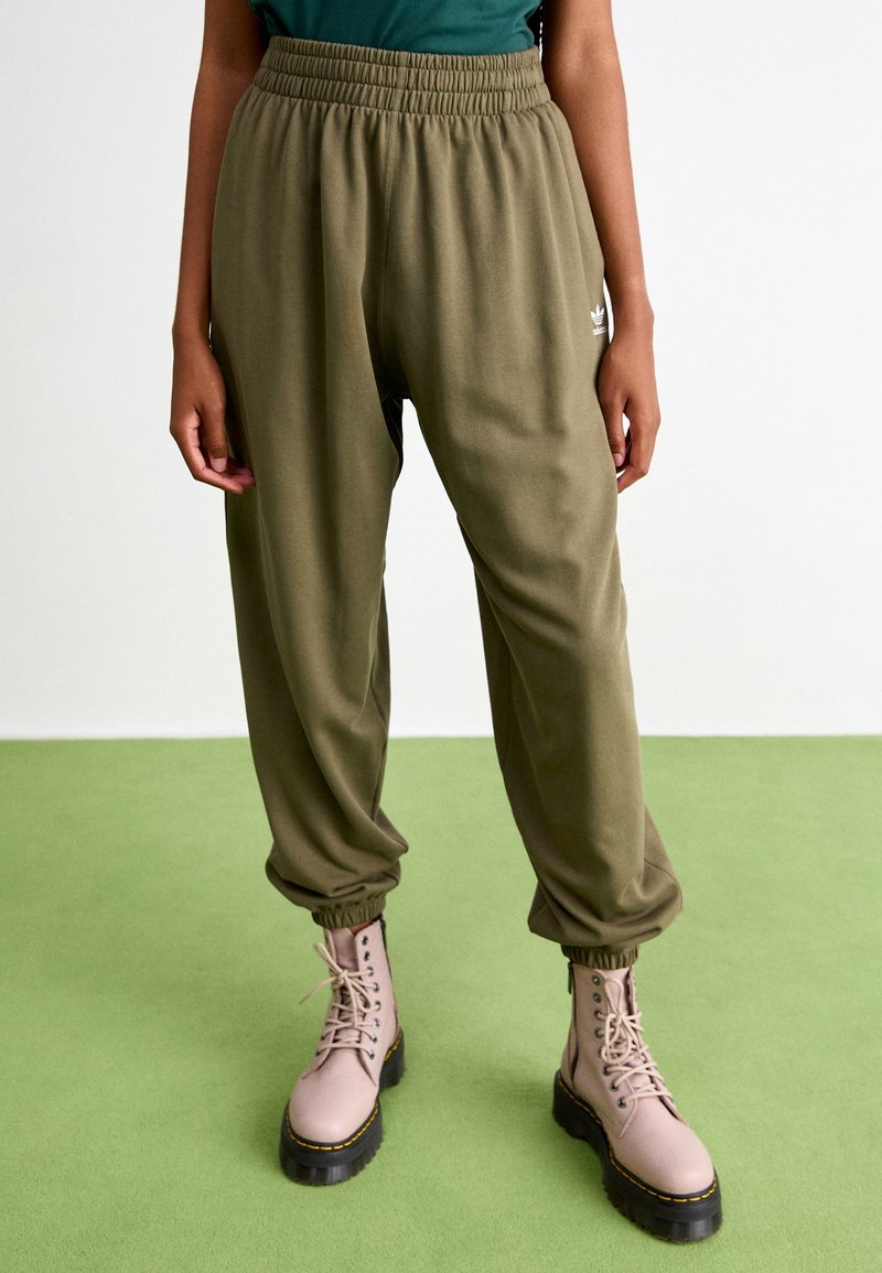 Osoba oblečená v olivově zelených Adidas teplácích a světle růžových šněrovacích botách, stojící na zelené podlaze s bílým pozadím.