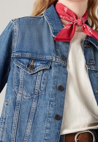 Jeansjacka i blekt blått med bruna knappar, två bröstfickor och en spetsig krage, kombinerad med en röd bandana runt halsen.