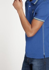 Kortärmad polo skjorta i livlig blå, med en treknapplåsning, kontrasterande krage och ärmkanter samt subtil detalj med logotyp.