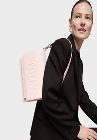 Rosa Leder-Rucksack mit geprägtem "TOUS"-Logo, strukturierter Formgebung und einer mit Ketten verzierten Tragegurt, getragen über einem schwarzen Blazer.
