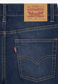 Jeans de mezclilla azul oscuro con un diseño de costura amarilla, que presentan una etiqueta de cuero en la cintura y una pequeña etiqueta roja en el bolsillo trasero.