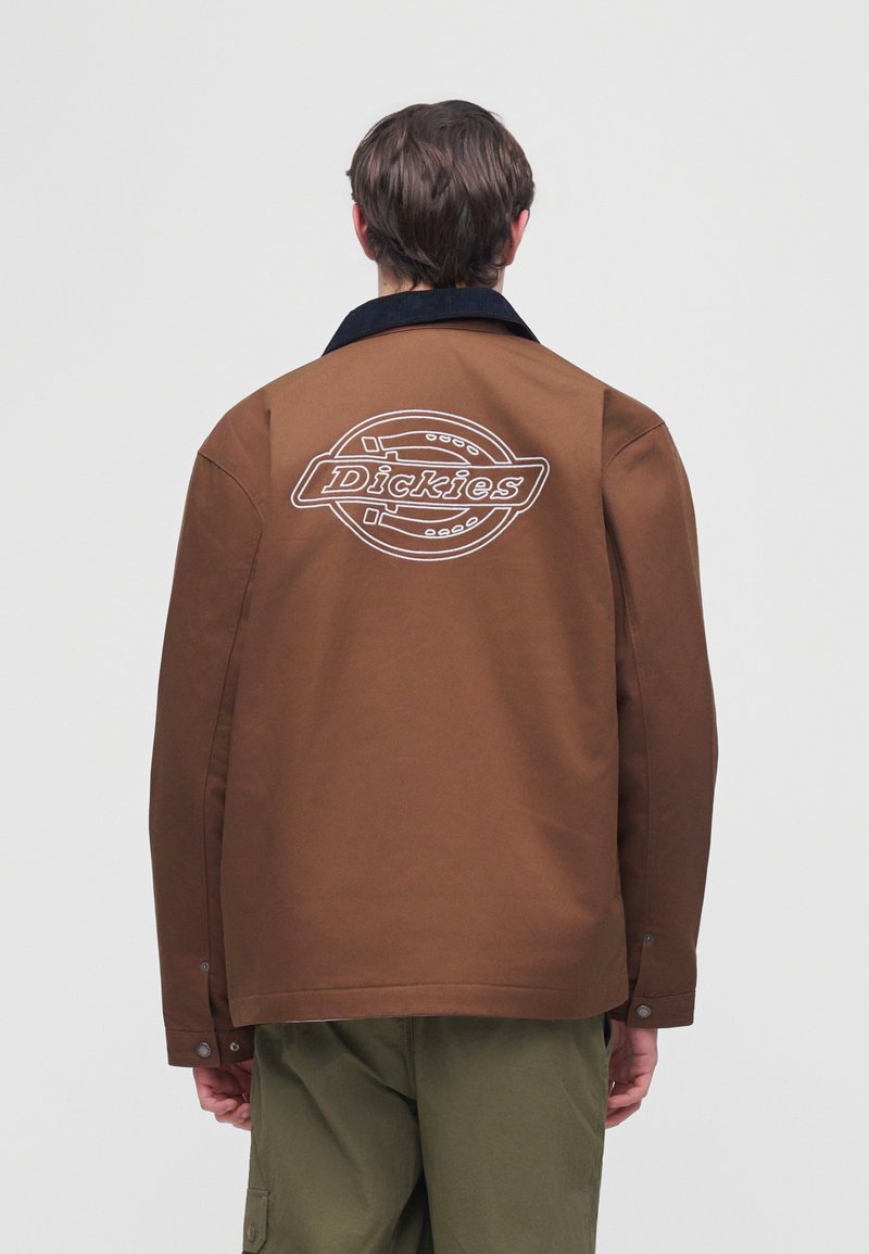 Person wearing brown jacket with black collar and large white Dickies logo on back, paired with olive green pants, facing away.