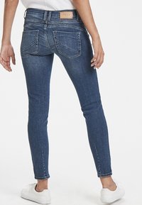 Jeans de denim azul ajustados con cintura alta, dos bolsillos traseros y un sutil desvanecimiento. Zapatillas blancas completan el look.