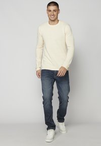 Beige texturiertes Langarmhemd mit gerippten Details, kombiniert mit dunkelblauen Jeans und weißen Turnschuhen. Das Model steht vor einem neutralen Hintergrund.