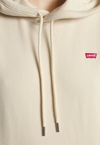 Sweat à capuche crème en tissu doux, avec une poche avant et des cordons tressés à embouts métalliques. Étiquette logo Levi's rouge sur la poitrine.