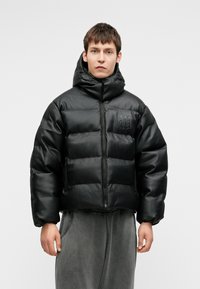 Črna napihnjena jakna z visokim ovratnikom, z zadrgo in embosiranim logotipom na prsih. Ima kvadratno silhueto in elastične manšete.