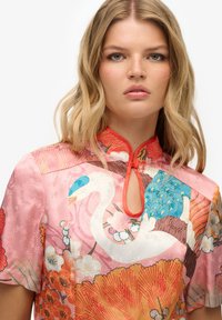 Blusa rosa a maniche corte con motivi floreali e di uccelli, dotata di un collo a chiave rifinito in rosso. Tessuto morbido con una leggera lucentezza.