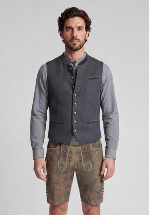 Mann in traditioneller bayerischer Tracht mit grauem Hemd, dunkelgrauer Knopfweste und braunen bestickten Lederhosen vor neutralem Hintergrund.