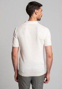 Mężczyzna odwrócony plecami, w krótkim rękawie białej pikowanej koszulce polo i szarych spodniach, na jednolitym szarym tle.