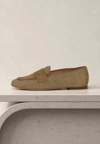 Μπεζ πλεκτό μοκασίνι με δερμάτινο λουράκι και στρίφωμα, τοποθετημένο σε ανοιχτή πέτρινη επιφάνεια με φόντο ανοιχτό χρώμα.