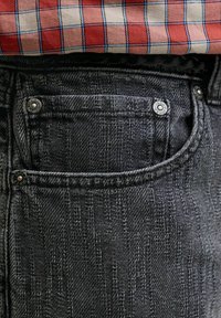 Detailný záber na čiernu štruktúrovanú rifľovú kapsu so striebornými nitmi pod lemom červeno-béžovej kockovanej košele.