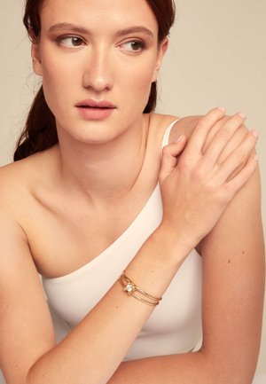 Bracelet en or avec plusieurs bandes entrelacées, comportant une grande pierre précieuse centrale. Le mannequin présente le bracelet sur une épaule dénudée.