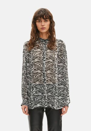 Kobieta o kręconych włosach, ubrana w przezroczystą bluzkę z nadrukiem zebry z długim rękawem i czarne skórzane spodnie, stojąca na jednolitym tle.
