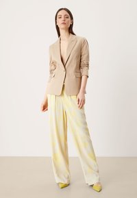 Beige blazer met een enkele knop en opgerolde mouwen, gecombineerd met luchtige gele patroonbroek en gele puntige hakken.