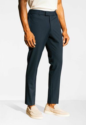 Homme portant un pantalon slim bleu marine foncé et des chaussures beige à enfiler, debout, les mains détendues le long du corps, sur un fond uni.