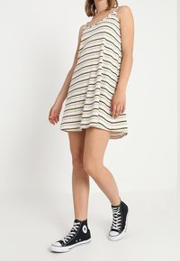 Mujer vestida con un vestido sin mangas de corte A a rayas y zapatillas negras de caña alta, de pie frente a un fondo blanco liso.