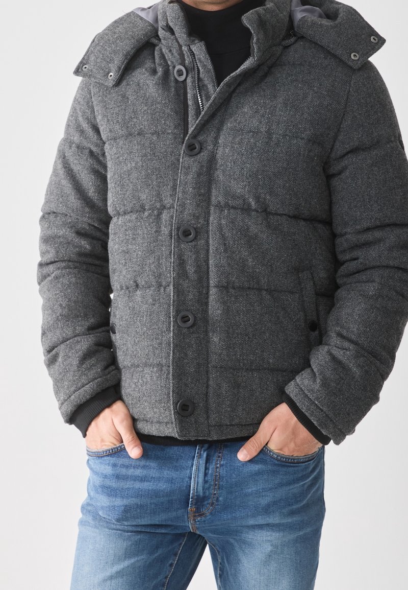 Mann trägt eine graue gesteppte Jacke mit Knöpfen, steht mit den Händen in den Taschen, kombiniert mit blauen Jeans und sichtbaren schwarzen Innensäumen.