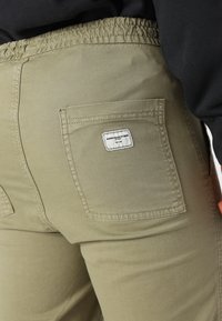 Olivgröna byxor i stretchmaterial med elastisk midja och en ficka på baksidan som har en liten tygetikett. Slät textur.