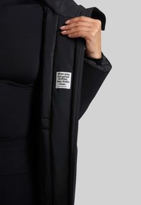 Sort polstret overtøj med en glat tekstur, der har et synligt indvendigt mærke med en sætning og hjemmeside. Designet inkluderer en bred åbning.