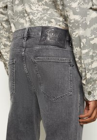 Gray denim jeans with a textured finish. Features a leather brand patch, two back pockets, and a straight cut design. Patterned shirt partially visible.