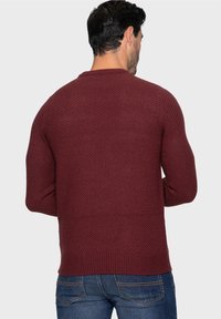 Burgunderroter Strickpullover mit strukturiertem Muster, Rundhalsausschnitt und gerippten Bündchen, kombiniert mit dunkelblauen Jeans. Sichtbare Rückansicht.