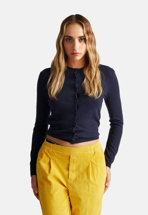 Marineblauwe lange mouwen cardigan met een ronde hals en knopen aan de voorkant, gecombineerd met felgele corduroy broek.
