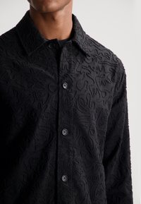 Svart skjorte med et teksturert mønster, knappelukking og en tradisjonell krage. Stoffet har et subtilt hevet, abstrakt design.