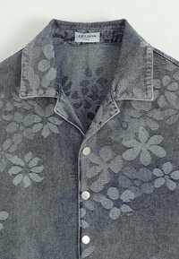 Chemise en denim gris foncé avec motif floral, dotée d'un col pointu et de boutons argentés sur le devant. Texture lisse, design décontracté.