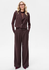 Brązowa dopasowana kamizelka i spodnie o szerokich nogawkach, zestawione z przezroczystą bluzką z długim rękawem. Charakteryzuje się dużymi klapami i dwoma bocznymi kieszeniami. Błyszczące szpilki.