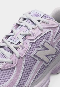 Κοντινό πλάνο ενός μωβ αθλητικού παπουτσιού New Balance με λευκά κορδόνια και ύφασμα από δίχτυ, που δείχνει το λογότυπο "N" της μάρκας στο πλάι.