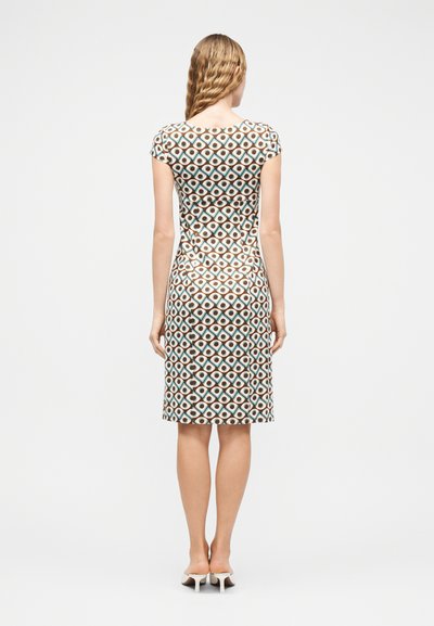 Γυναίκα με κυματιστά ξανθά μαλλιά, φοράει εφαρμοστό φόρεμα μήκους μέχρι το γόνατο με γεωμετρικό μοτίβο σε καφέ, τιρκουάζ και λευκά σχήματα, στέκεται γυρισμένη προς τα πίσω.